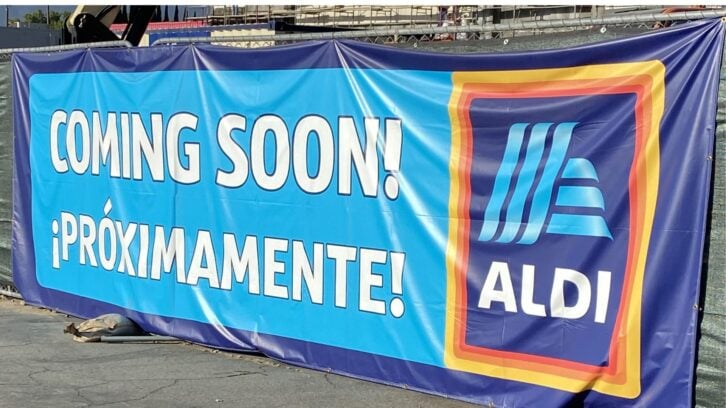A blue banner on a fence displays the words "COMING SOON! ¡PRÓXIMAMENTE!" next to the ALDI supermarket logo.