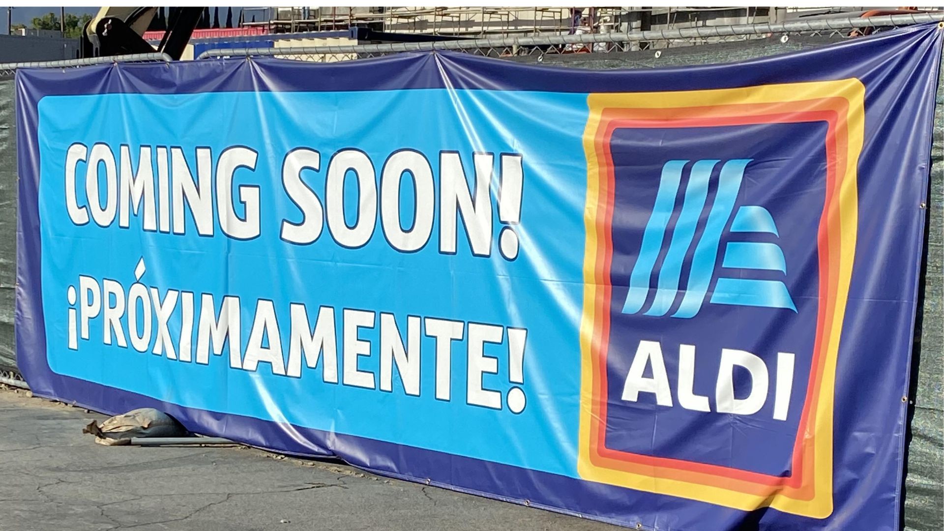 A blue banner on a fence displays the words "COMING SOON! ¡PRÓXIMAMENTE!" next to the ALDI supermarket logo.