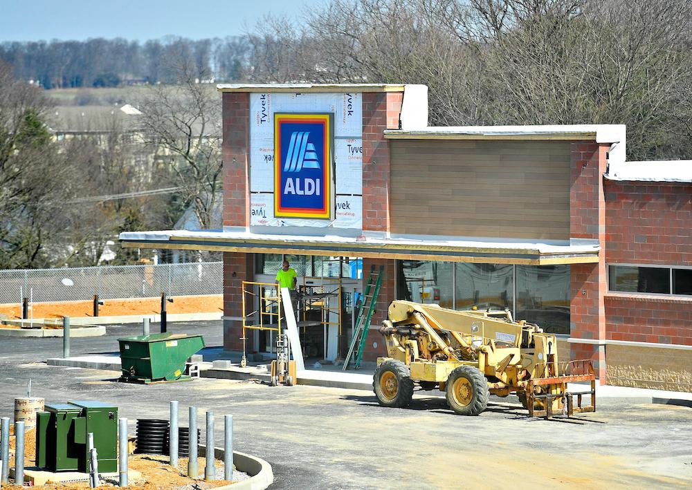 A worker stands on a scaffold outside an ALDI store under construction, with building materials and a forklift visible in the parking lot. Trees and fields are in the background.
