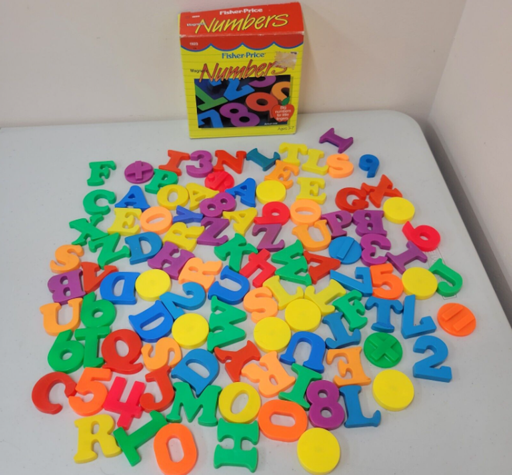 Colorful plastic letters and numbers are scattered on a white table, with a yellow Fisher-Price Numbers box in the background. The pieces are in various colors, including red, green, yellow, orange, and blue.