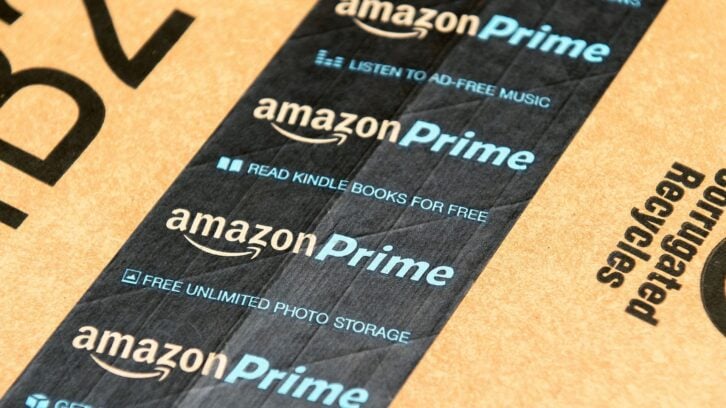 A cardboard shipping box with Amazon Prime-branded tape, promoting ad-free music, free Kindle books, and unlimited photo storage, along with the Amazon Prime logo.