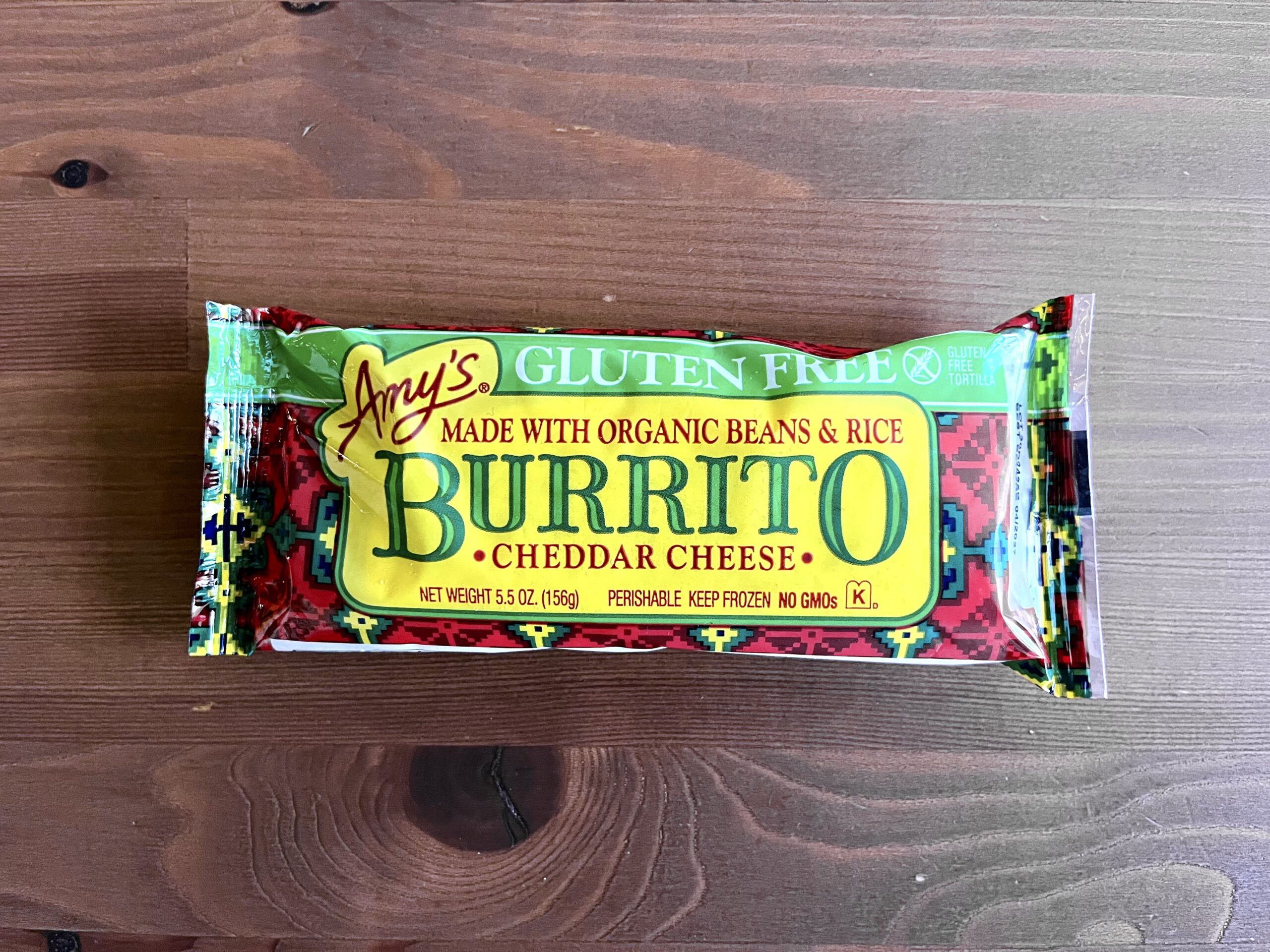 A packaged Amy’s gluten-free burrito with organic beans, rice, and cheddar cheese lies on a wooden surface. The wrapper is colorful with yellow and red accents.