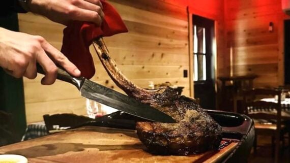 A person slices one of the biggest steaks—a large, cooked tomahawk steak—on a wooden cutting board in a rustic, warmly lit restaurant with wooden walls and empty chairs in the background.