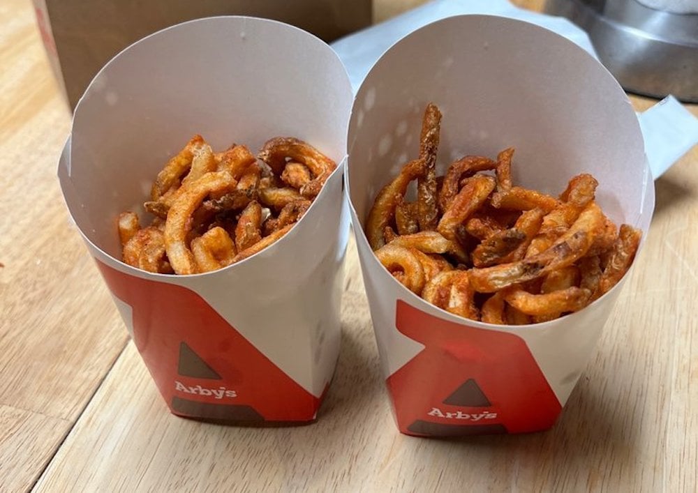 Two containers of Arby’s seasoned curly fries are placed side by side on a wooden surface. The fries are golden brown and spilling slightly over the tops of the white and red paper containers.