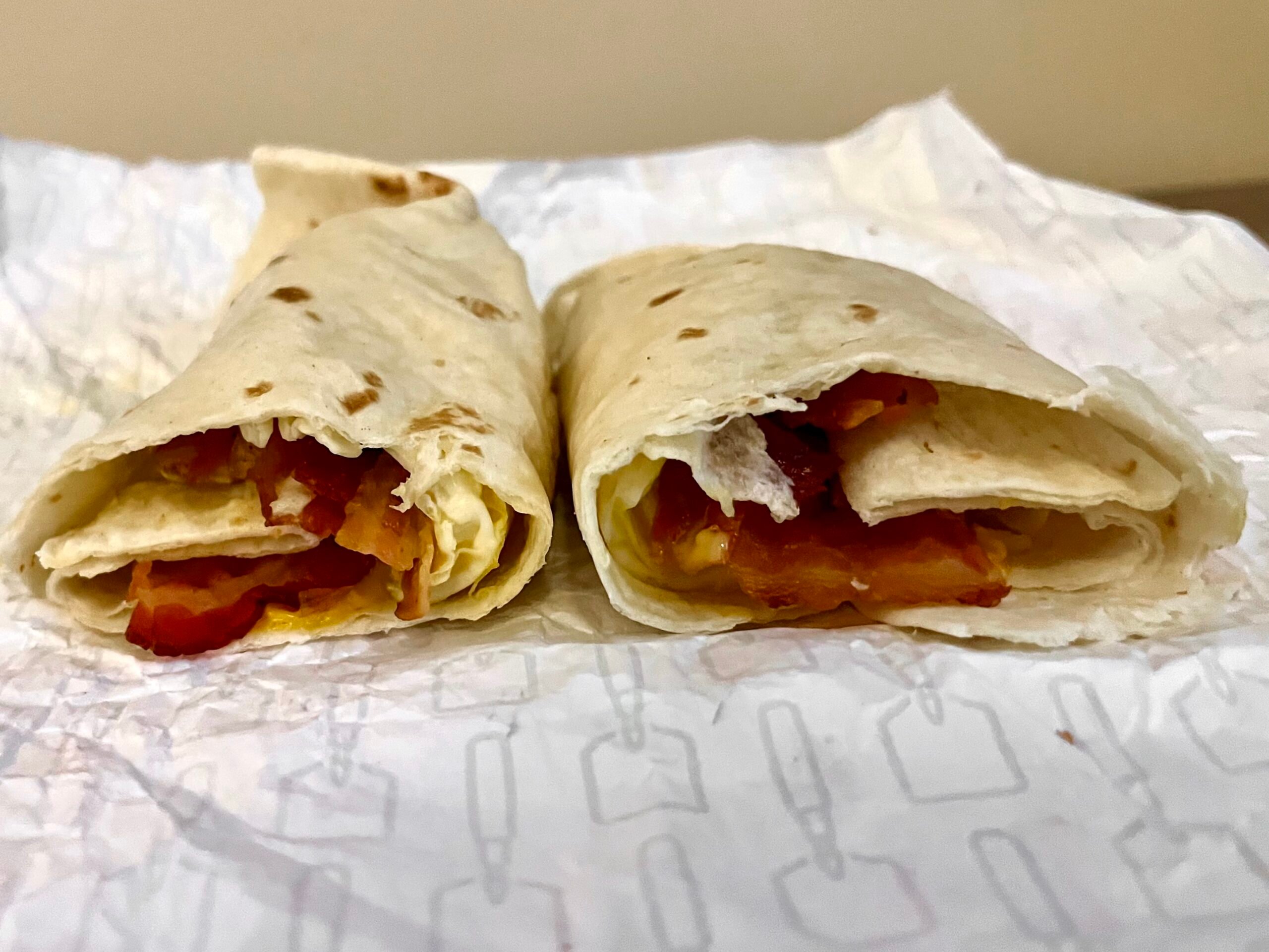 Two breakfast wraps filled with bacon and scrambled eggs sit on crinkled paper, cut open to reveal the fillings inside.