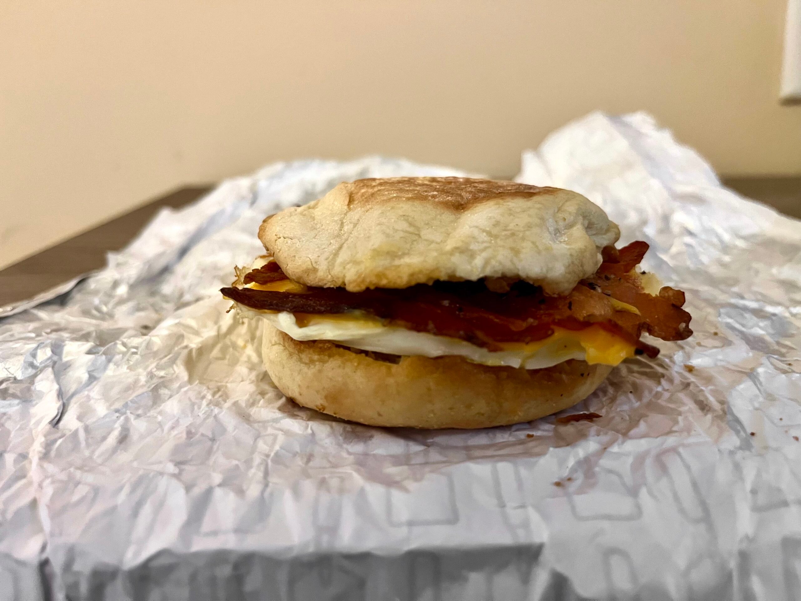 A breakfast sandwich with egg, bacon, and cheese on an English muffin sits on crinkled white sandwich paper, placed on a table with a plain beige background.