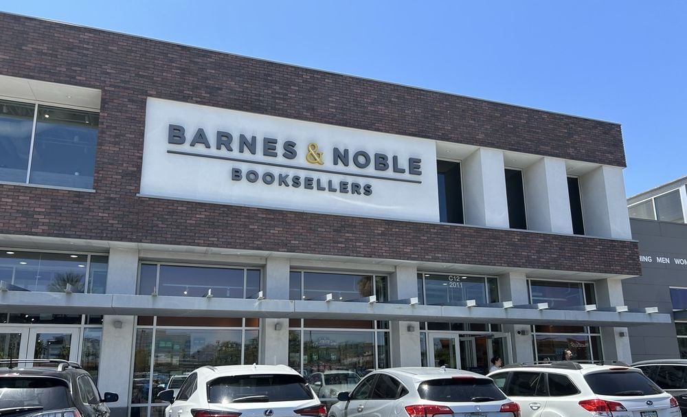 The image shows the exterior of a Barnes & Noble Booksellers store, with several parked cars in front under a clear blue sky. The store sign is prominently displayed above the entrance.