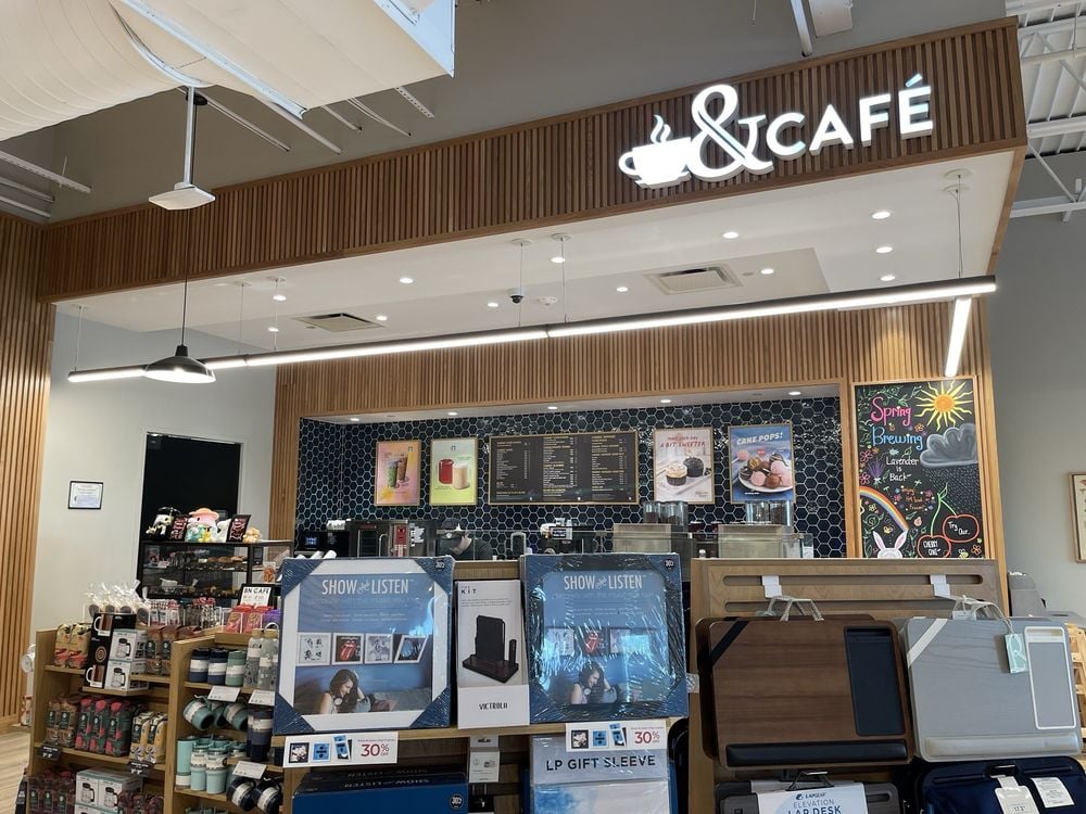 A modern café counter with a stylish wood and black tile design, digital menu boards, and various products for sale, including coffee makers, mugs, and vinyl records display racks in the foreground.