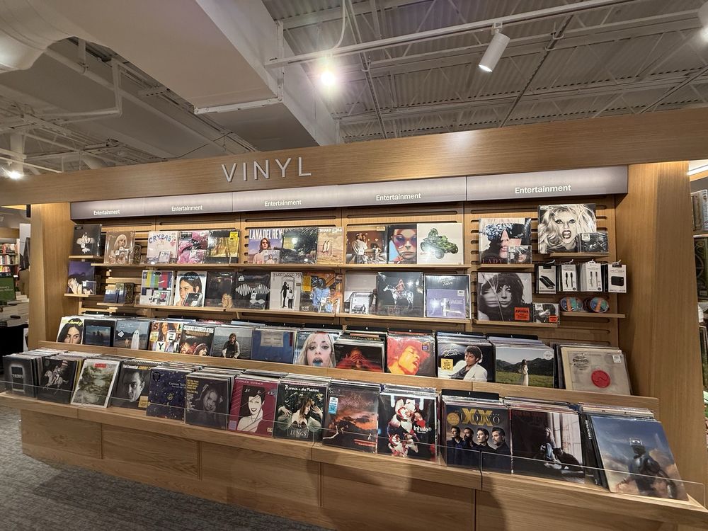 A well-lit store display labeled "Vinyl" features multiple shelves filled with assorted vinyl records, each showing colorful album covers from various artists and genres. The shelves are organized and neatly arranged.