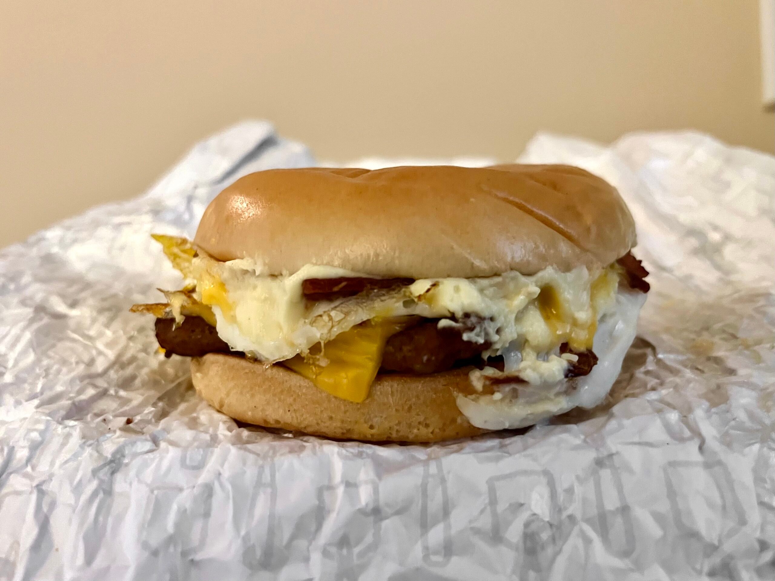 A breakfast sandwich with egg, melted cheese, and meat on a bun, sitting on crumpled white paper against a plain background.