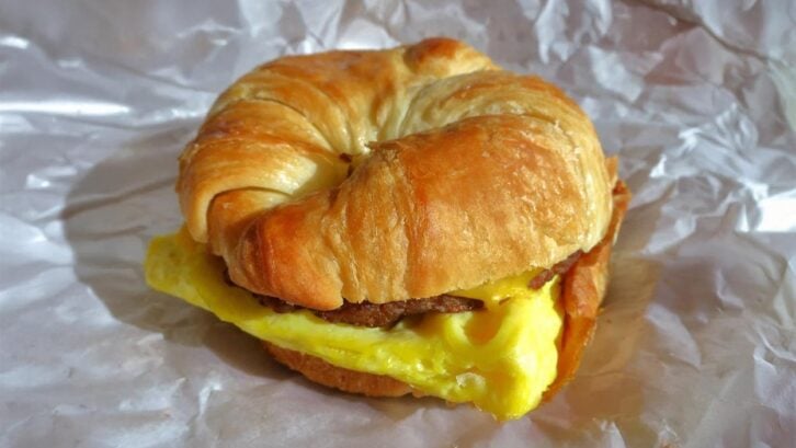A croissant breakfast sandwich, inspired by classic fast-food breakfast sandwiches, filled with a folded egg, sausage patty, and melted cheese, served on crinkled white paper.