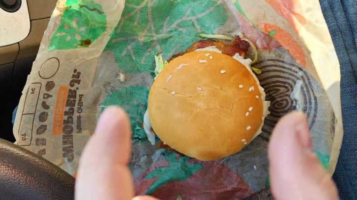 A sesame seed hamburger on fast food wrapper sits on someone's lap in a car, with two fingers visible in the foreground about to grab the burger.