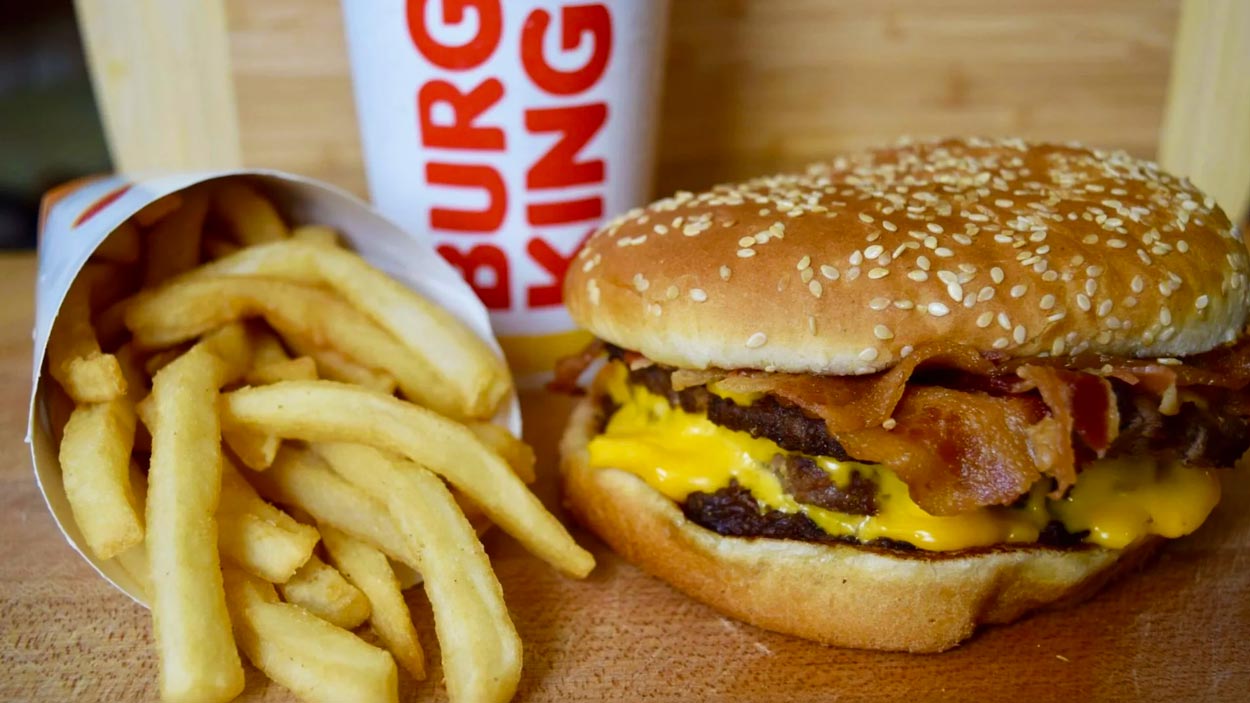 A Burger King meal with a bacon cheeseburger, a serving of French fries in a branded container, and a soft drink cup displaying the Burger King logo.