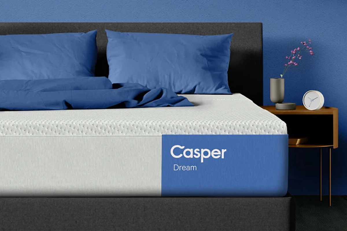 A neatly made bed with a gray headboard, blue sheets and pillows, and a white mattress labeled "Casper Dream" in a blue rectangle. A small wooden nightstand holds a vase with flowers, a book, and a white clock.