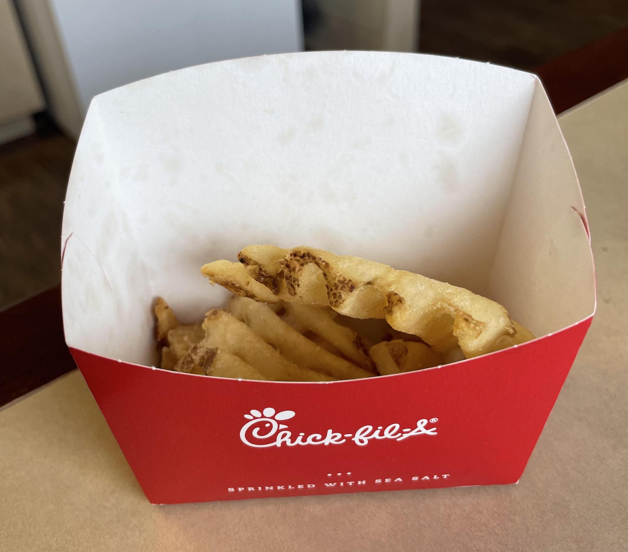 A red Chick-fil-A container holding a few waffle fries sits on a beige tabletop. The fries appear to be mostly eaten, with only a few pieces left in the box.