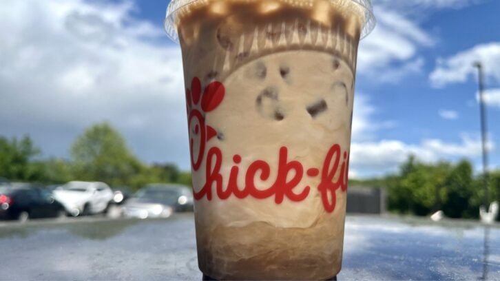 A plastic Chick-fil-A cup filled with iced coffee sits on a reflective surface outdoors, with a cloudy blue sky and parked cars in the background.