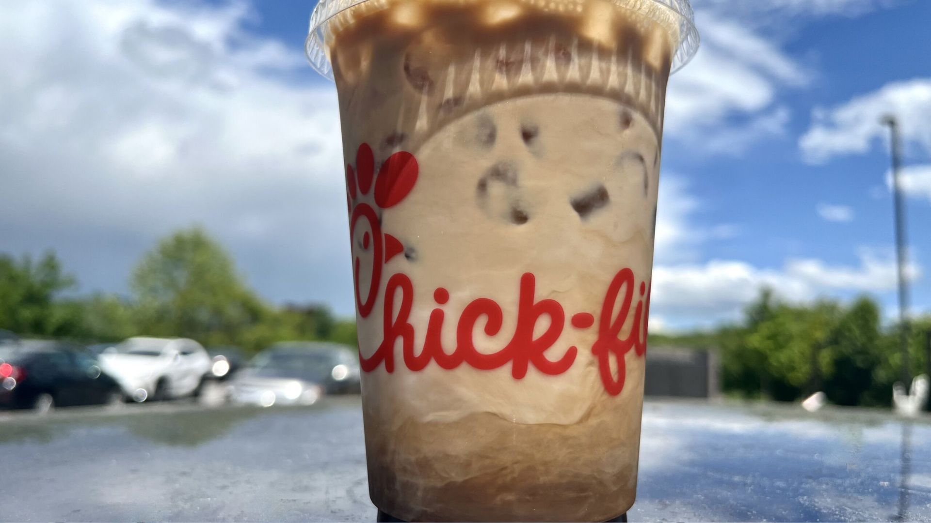 A plastic Chick-fil-A cup filled with iced coffee sits on a reflective surface outdoors, with a cloudy blue sky and parked cars in the background.