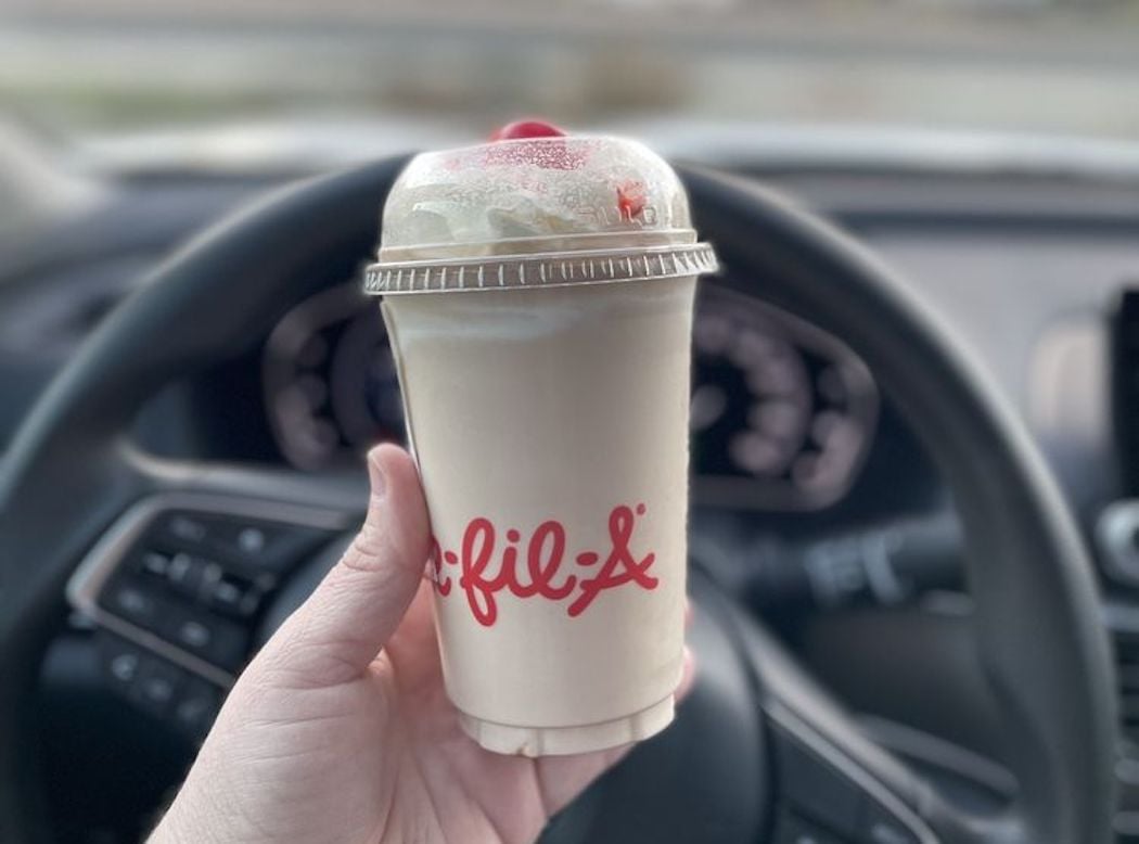 A hand holds a Chick-fil-A milkshake inside a car, with the dashboard and steering wheel visible in the background.