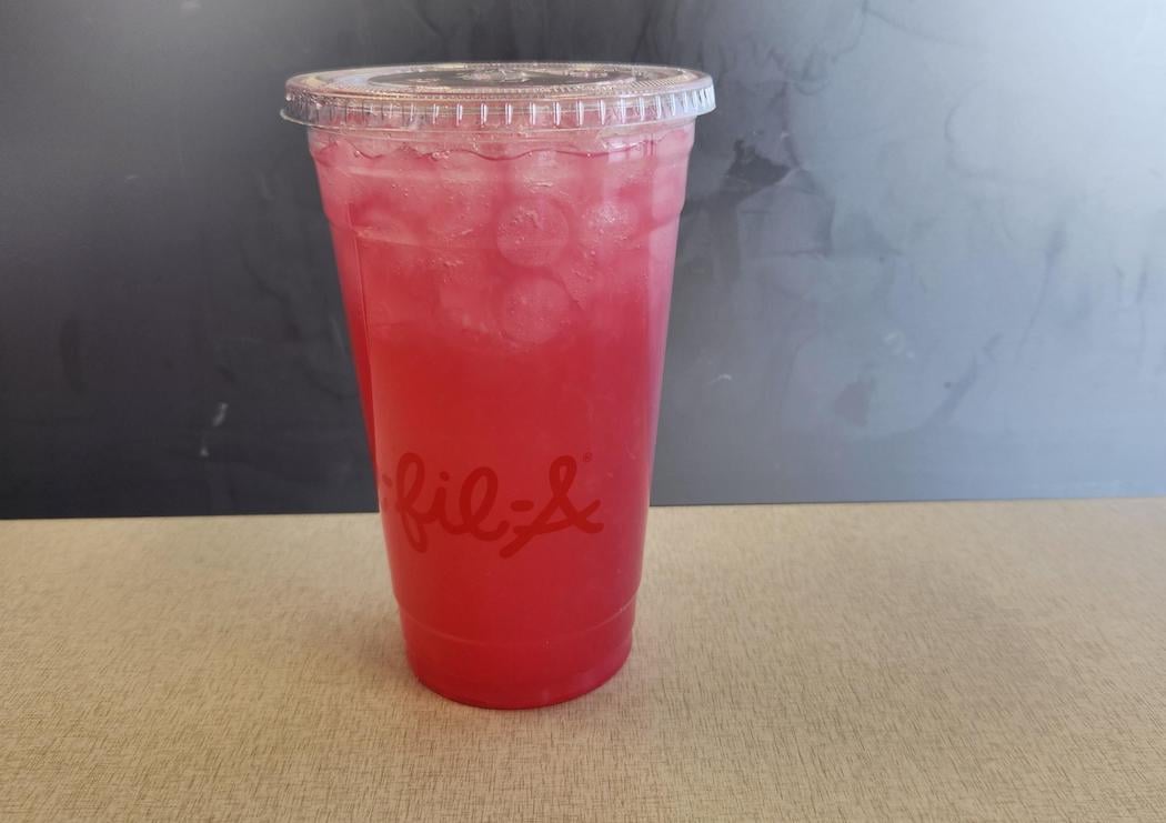 A Pineapple Dragonfruit Lemonade clear plastic cup with a lid, filled with pink iced lemonade, sits on a light-colored tabletop. The cup has the red Chick-fil-A logo printed on it. The background is dark and plain.