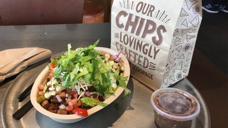 A bowl of Mexican-style food featuring chipotle's carne asada is topped with lettuce, corn, cheese, and salsa, served on a tray beside a bag labeled “Our Chips Are Lovingly Crafted” and a small cup of salsa with a napkin and black fork.