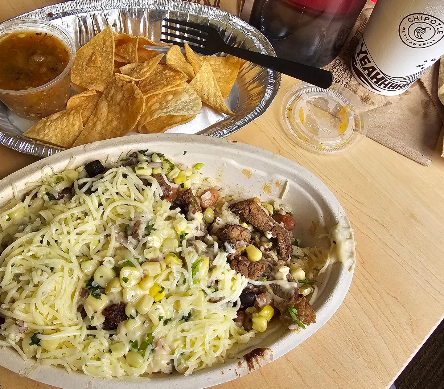 A burrito bowl with cheese, corn, beans, and steak is next to a tray of tortilla chips with salsa, a plastic fork, a drink cup, and a napkin on a wooden table. Chipotle bowl