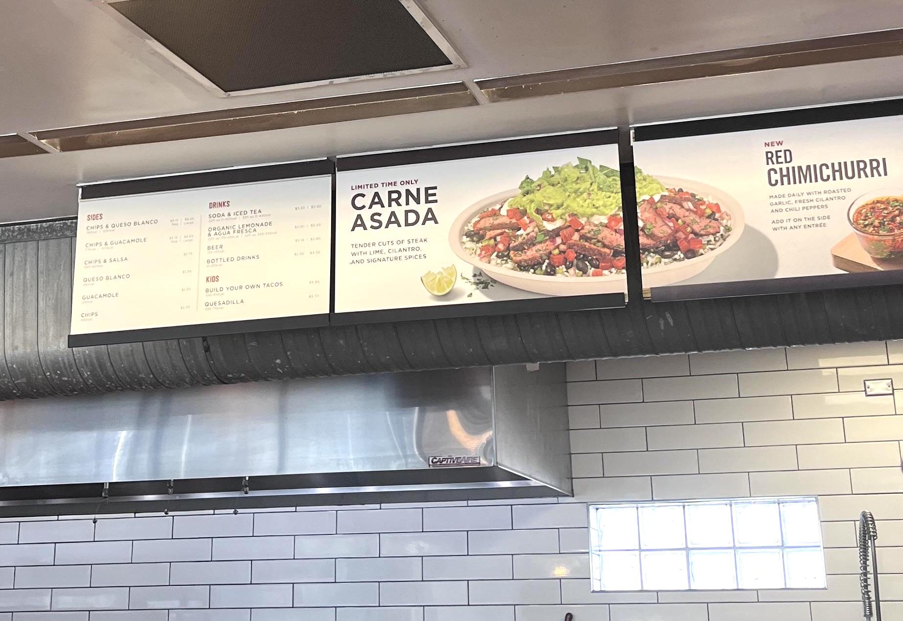 Menu board inside a Chipotle restaurant featuring a "Carne Asada" bowl with steak, salsa, and guacamole, plus a new "Red Chimichurri" dish. Tacos, burritos, and drinks are also listed on the left. White tile background.