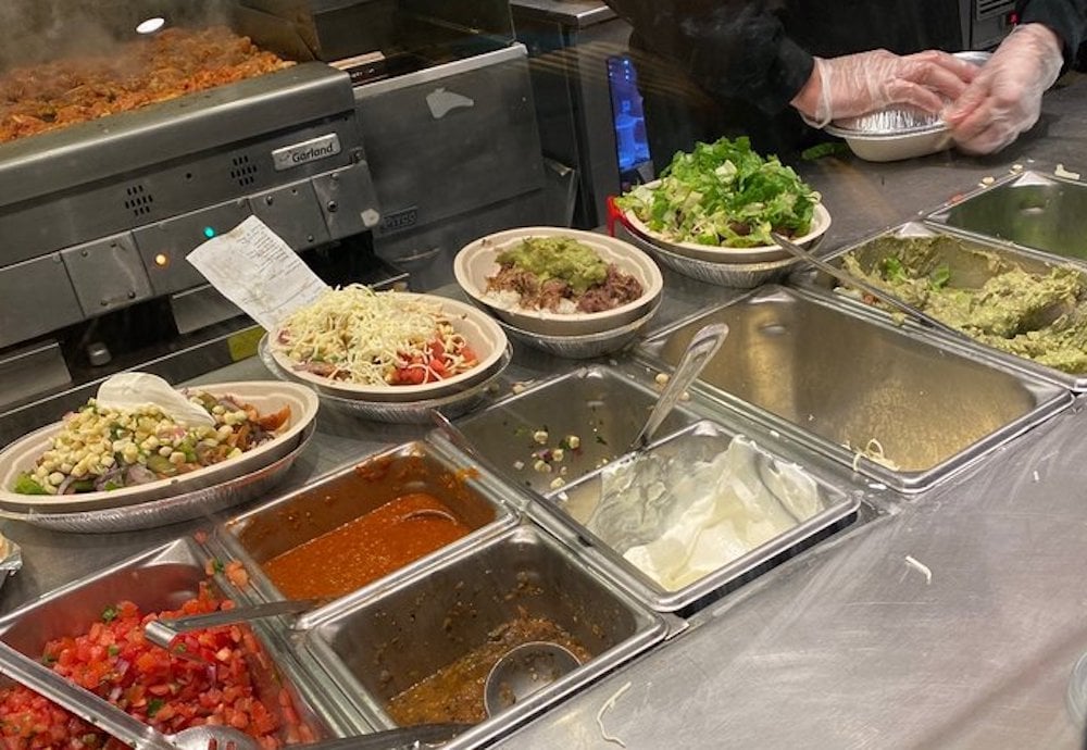A Chipotle server wearing gloves prepares burrito bowls behind a counter filled with fresh toppings, including salsa, beans, cheese, lettuce, and guacamole, at a Chipotle fast-casual restaurant.