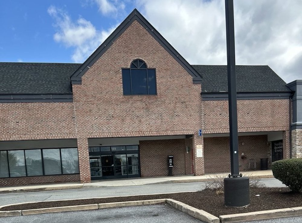A vacant brick storefront with glass doors and windows, an arched window above, and a small mailbox near the entrance. The parking lot in front is mostly empty, with a tall lamp post and some landscaping visible.