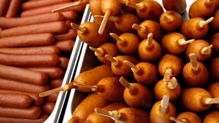 A close-up view of plain hot dogs lined up on the left and a pile of golden-brown corn dogs with wooden sticks on the right in metal trays, offering a tasty snack selection despite recent news of a corn dog recall.
