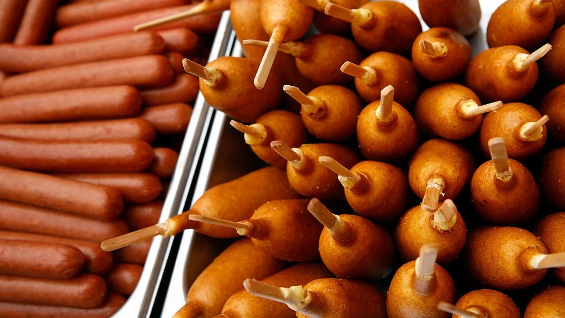 A close-up view of plain hot dogs lined up on the left and a pile of golden-brown corn dogs with wooden sticks on the right in metal trays, offering a tasty snack selection despite recent news of a corn dog recall.