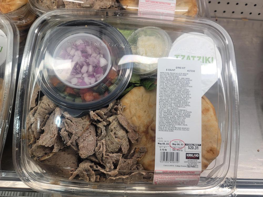 Plastic container with gyro meat slices, pita bread, tzatziki sauce, a small cup of diced red onions and tomatoes, and a clear lid with a printed label showing price and ingredients.
