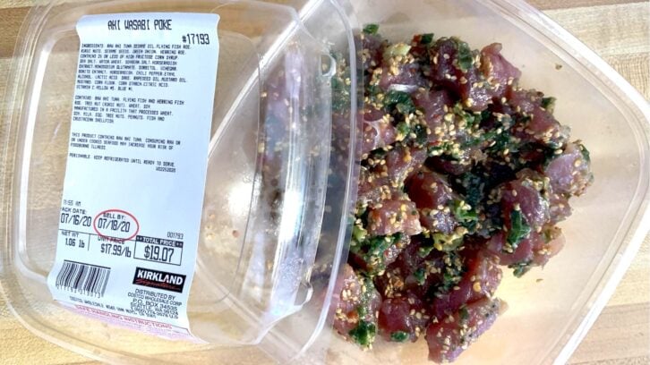 A plastic container of Kirkland Ahi Wasabi Poke with a clear lid, showing chunks of marinated fish mixed with green onions and sesame seeds. The product label and price are visible on the left.