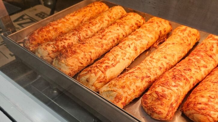 Several golden-brown cheese-topped breadsticks are lined up on a metal tray inside a display case, with a crispy and slightly melted cheese coating on top.