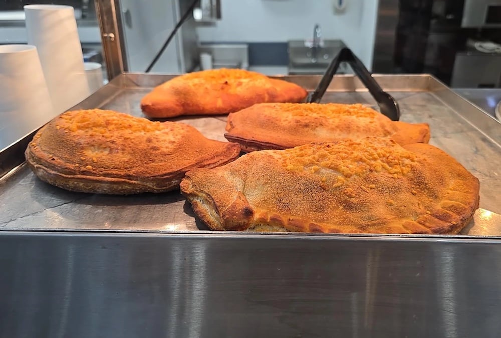 Four golden-brown Costco food court calzones with crispy, textured crusts are displayed on a metal tray in a food service setting. Black tongs rest on the tray behind the calzones.