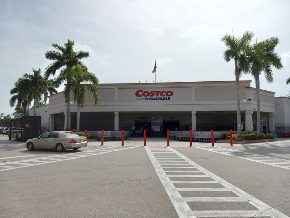 A Costco Wholesale store with palm trees around the parking lot, a few cars parked in front, and a clear sky overhead. Red bollards line the entrance.