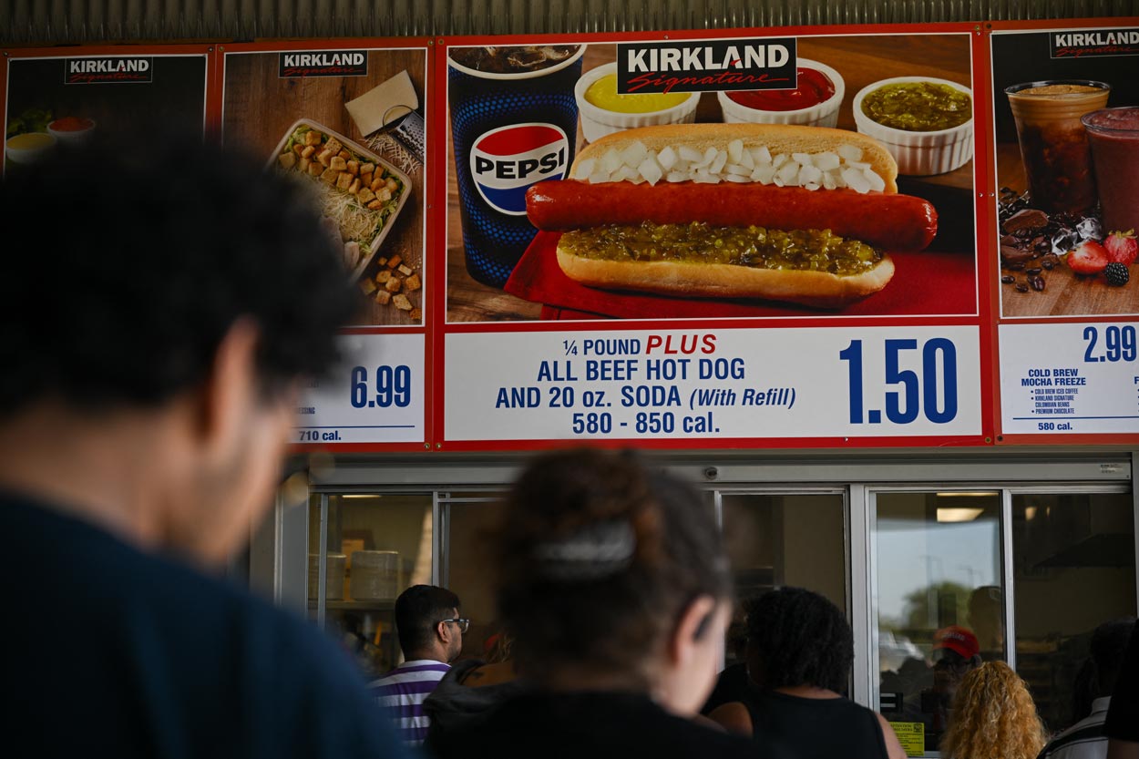 People stand in line beneath a sign advertising a $1.50 combo meal of a 1/4 pound all beef hot dog with soda at Costco’s food court—one of the classic Costco products loved by shoppers, featured prominently on the sign.