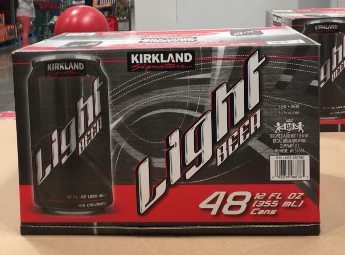 A 48-pack of Costco Kirkland Signature Light Beer cans sits on a store shelf. The packaging is black and red, with "Light Beer" in large white letters and "48 12 FL OZ (355 mL) Cans" in bold text on the front.