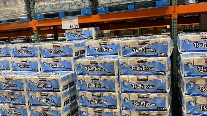 Stacks of Kirkland Signature Bath Tissue packages are displayed on metal shelves in a Costco warehouse store. A price sign above shows $16.99. The packages are mostly blue and white.