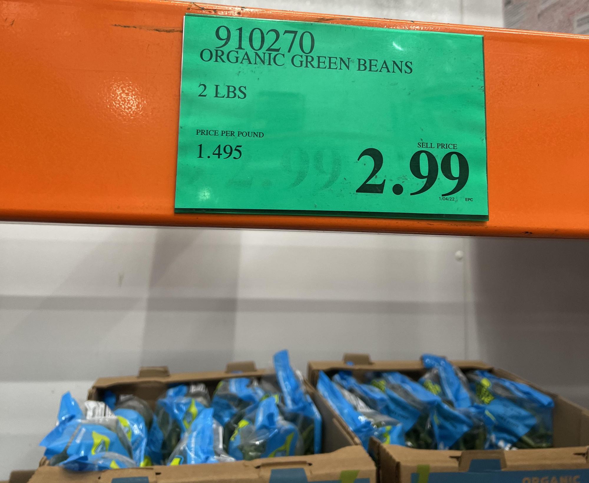 A store shelf displays boxes of organic green beans in blue packaging below a green price sign listing 2 pounds for $2.99, with a price per pound of $1.495 at Costco.