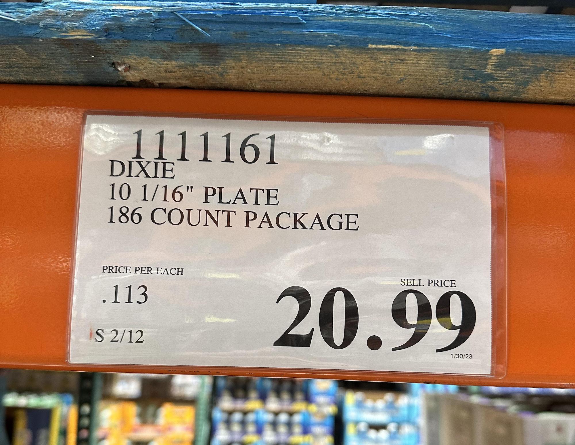 A store shelf label for Dixie 10 1/16-inch plates, 186 count package, showing a sell price of $20.99 and price per each plate as $0.113. Shelving and part of the Costco store are visible in the background.