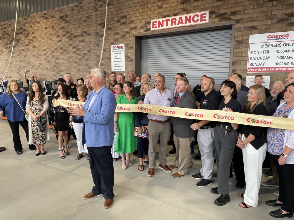 A group of people, including Costco staff and officials, stand in front of a Costco entrance for a ribbon-cutting ceremony. A man in a blue suit speaks while holding oversized scissors.