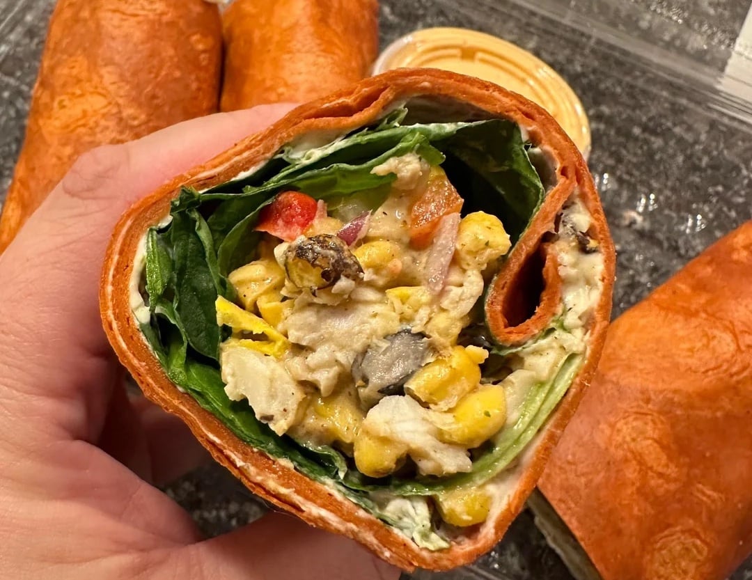A hand holds a tomato tortilla wrap filled with greens, corn, black beans, chopped vegetables, and a creamy dressing. More wraps and a container of orange dipping sauce are in the background.