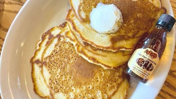 Three golden-brown pancakes topped with a pat of butter are served on a white plate next to a small bottle of pure maple syrup. The plate rests on a wooden table.