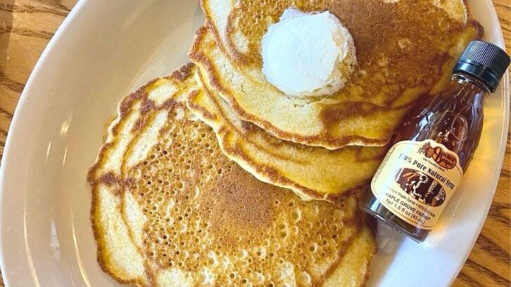 Three golden-brown pancakes topped with a pat of butter are served on a white plate next to a small bottle of pure maple syrup. The plate rests on a wooden table.