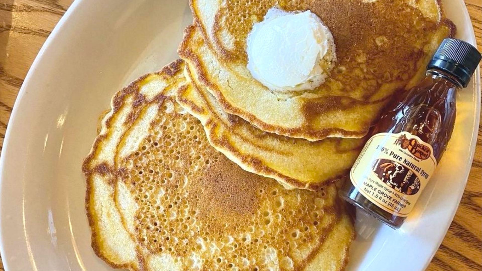 Three golden-brown pancakes topped with a pat of butter are served on a white plate next to a small bottle of pure maple syrup. The plate rests on a wooden table.