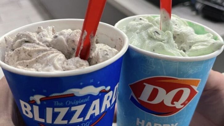 Two hands hold up Dairy Queen Blizzards: one in a blue cup with cookies and cream, the other in a blue cup with mint ice cream. Both have red spoons sticking out.