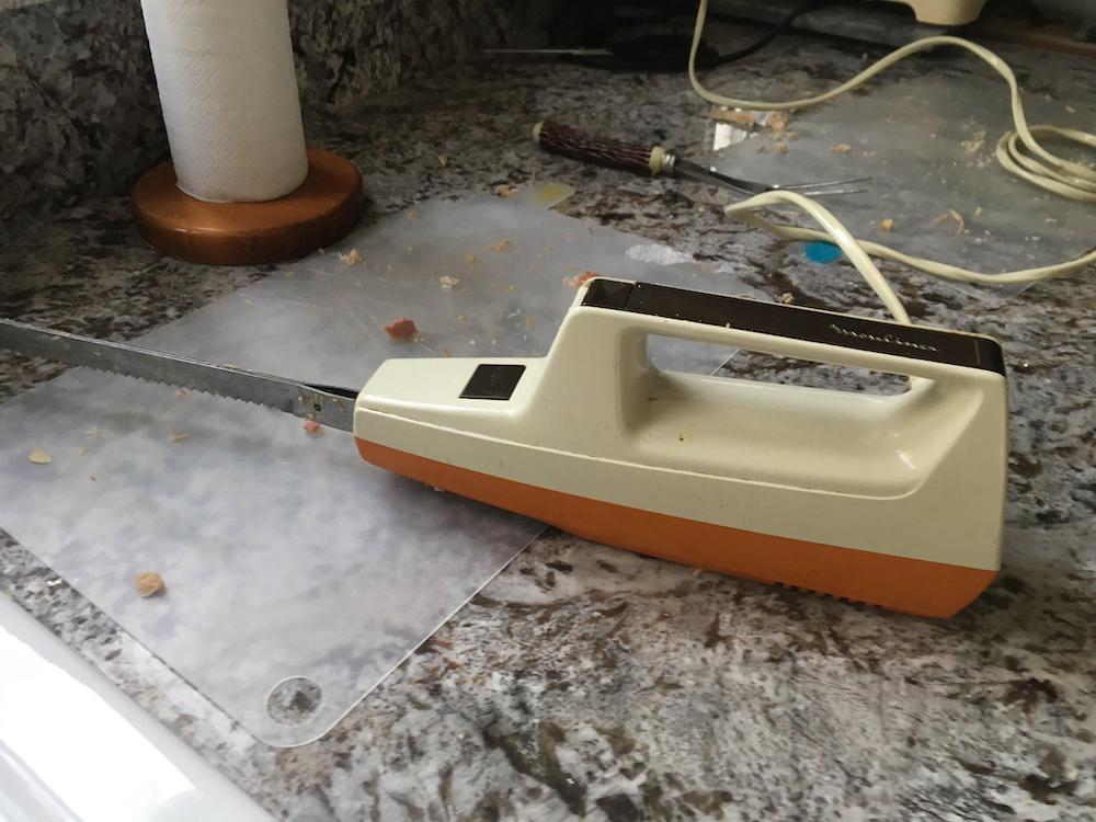 A 70s or 80s vintage electric knife with a white and orange handle rests on a messy kitchen counter with crumbs, a cutting board, a paper towel roll, and an unplugged cord in the background.