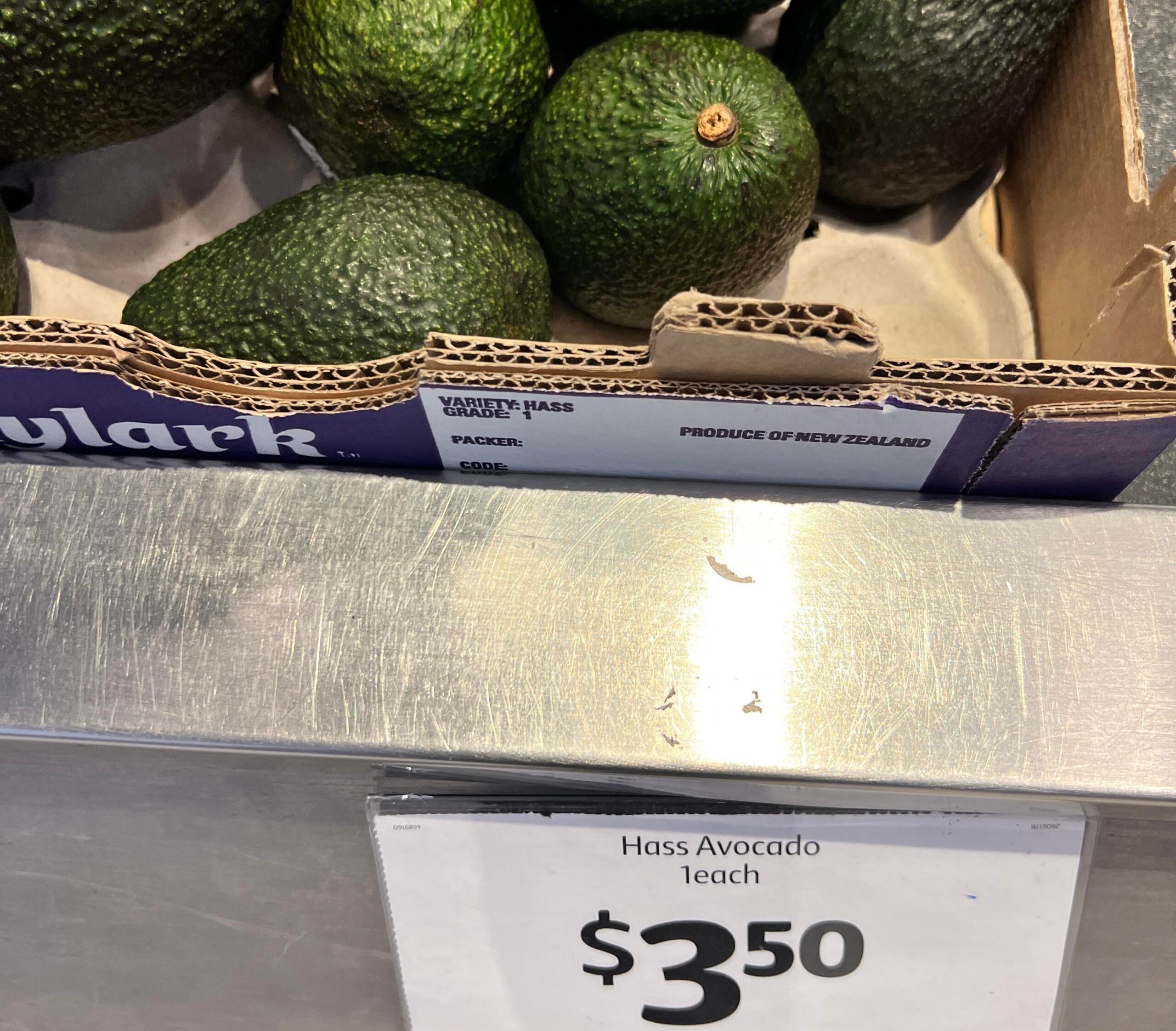 A box of Hass avocados from New Zealand is displayed in a grocery store, with a sign below reading "Hass Avocado each $3.50".