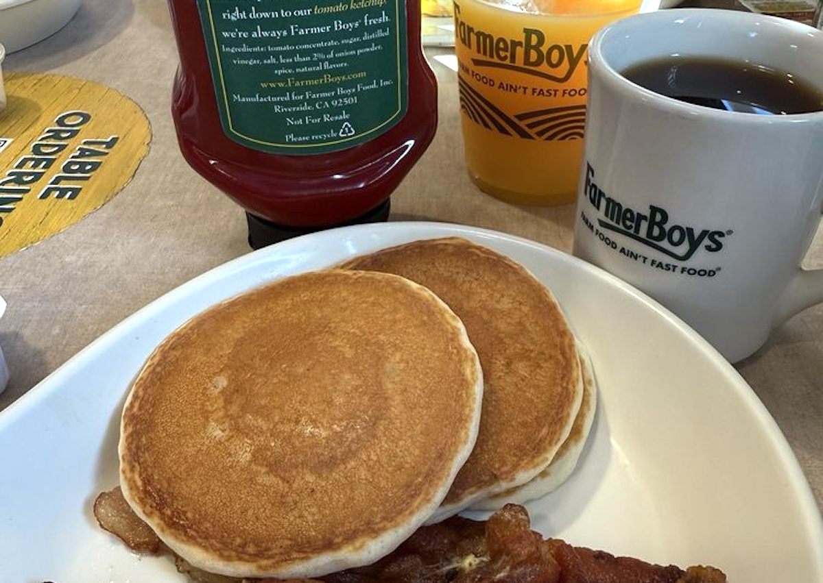 Two pancakes and strips of bacon on a white plate, with a cup of coffee, glass of orange juice, and ketchup bottle, all branded with Farmer Boys logos, set on a restaurant table.