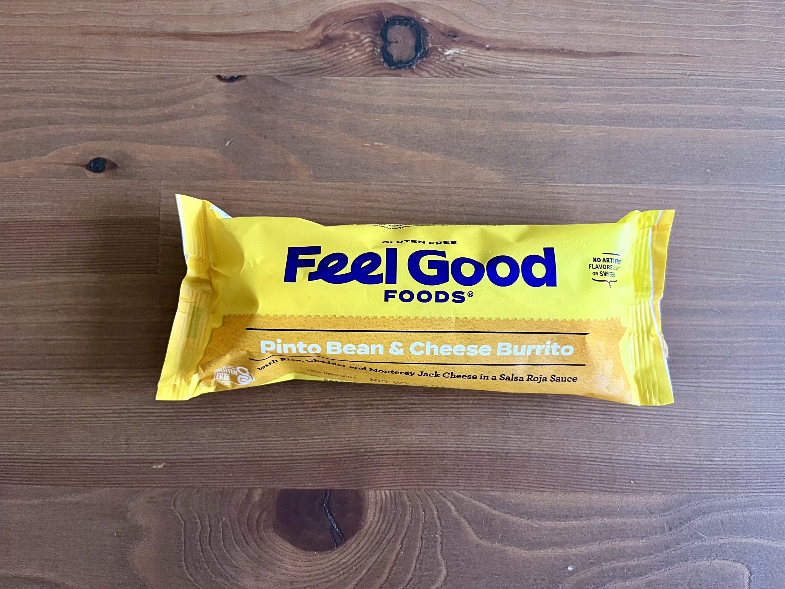 A yellow Feel Good Foods package of Pinto Bean & Cheese Burrito sits on a wooden surface. The packaging features bold text and details about the burrito's ingredients and gluten-free status.